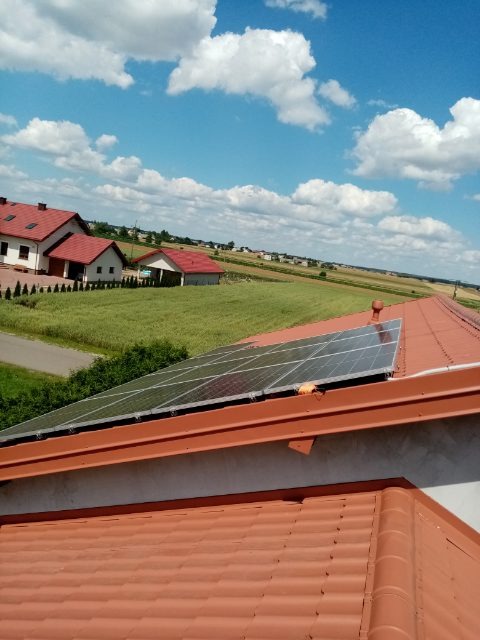 Panele fotowoltaiczne na czerwonym dachu domu jednorodzinnego, widok z góry na tle błękitnego nieba z chmurami i zielonego krajobrazu.