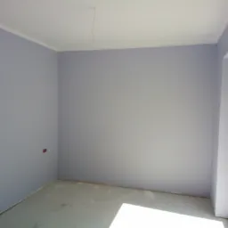 Świeżo pomalowane na fioletowo ściany w pustym pokoju z białym sufitem i listwami, widoczne ślady po remoncie na podłodze, światło słoneczne wpadające przez okno.