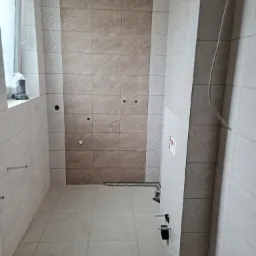 Wnętrze łazienki w trakcie prac wykończeniowych, widoczne ułożone płytki w dwóch odcieniach beżu, z przygotowanymi otworami na armaturę i instalacje, oraz okno z widocznym workiem z materiałem.