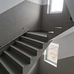 Klatka schodowa w budynku, ściany wykończone szarym tynkiem mozaikowym, szare schody z antypoślizgowymi rowkami, okna z widokiem na zewnątrz, poręcz zabezpieczona folią.