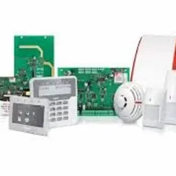 Zestaw komponentów systemu zabezpieczeń: centrala alarmowa z klawiaturą, czujnik dymu, czujnik ruchu i płytki drukowane elektroniki, na białym tle.