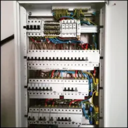 Wnętrze rozdzielnicy elektrycznej z widocznymi bezpiecznikami, przewodami i listwami zaciskowymi, zainstalowanej w białej obudowie.