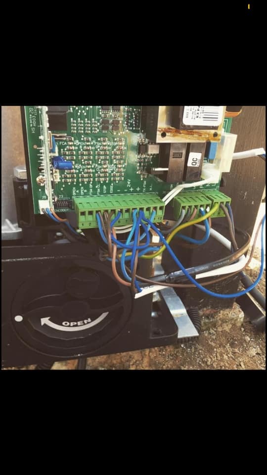 Zbliżenie na płytkę sterującą z zielonymi złączkami i podłączonymi przewodami w automatyce bramy, widoczny element obudowy z napisem 'OPEN' i strzałką.