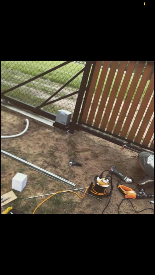 Montaż automatyki do przesuwnej bramy posesyjnej z drewnianymi panelami, widoczny napęd, przewody, wiertarka i szlifierka kątowa na trawie.