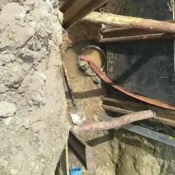 Głęboki wykop zabezpieczony drewnianymi belkami, widoczny pracownik w kasku i kraciastej koszuli w oddali, niebieskie rury i narzędzia w dole wykopu, woda zbierająca się na dnie.