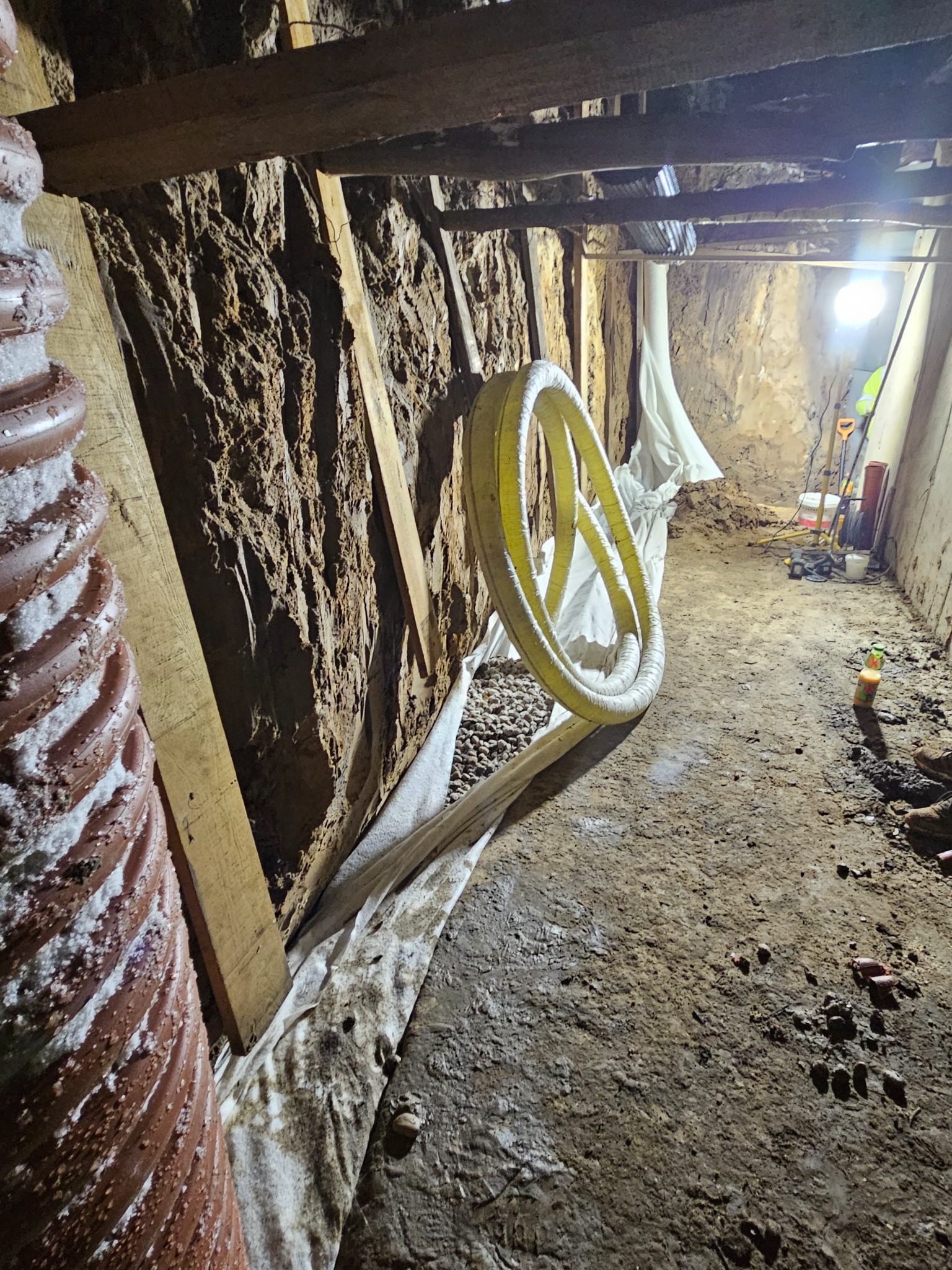 Wykop z odsłoniętą ziemią, zabezpieczony drewnianymi belkami, z widocznym żółtym wężem drenażowym i geowłókniną, oświetlony lampą roboczą.