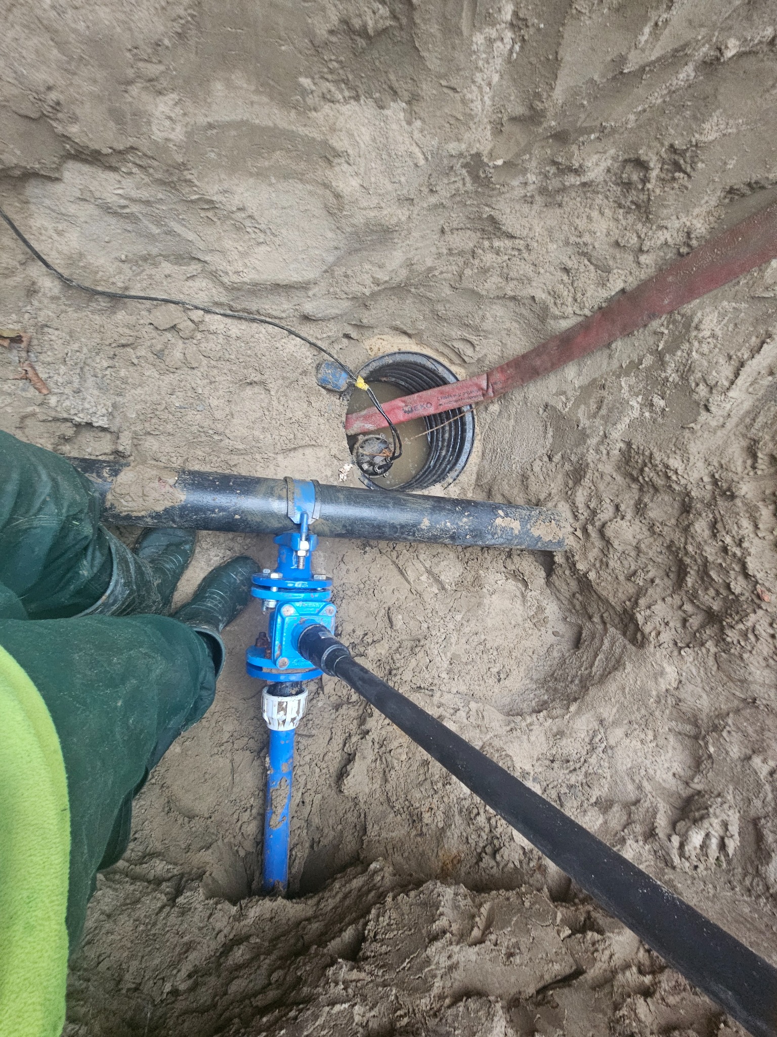 Niebieski zawór i czarne rury w wykopie ziemnym, widoczne fragmenty odzieży roboczej i studzienka kanalizacyjna w tle.