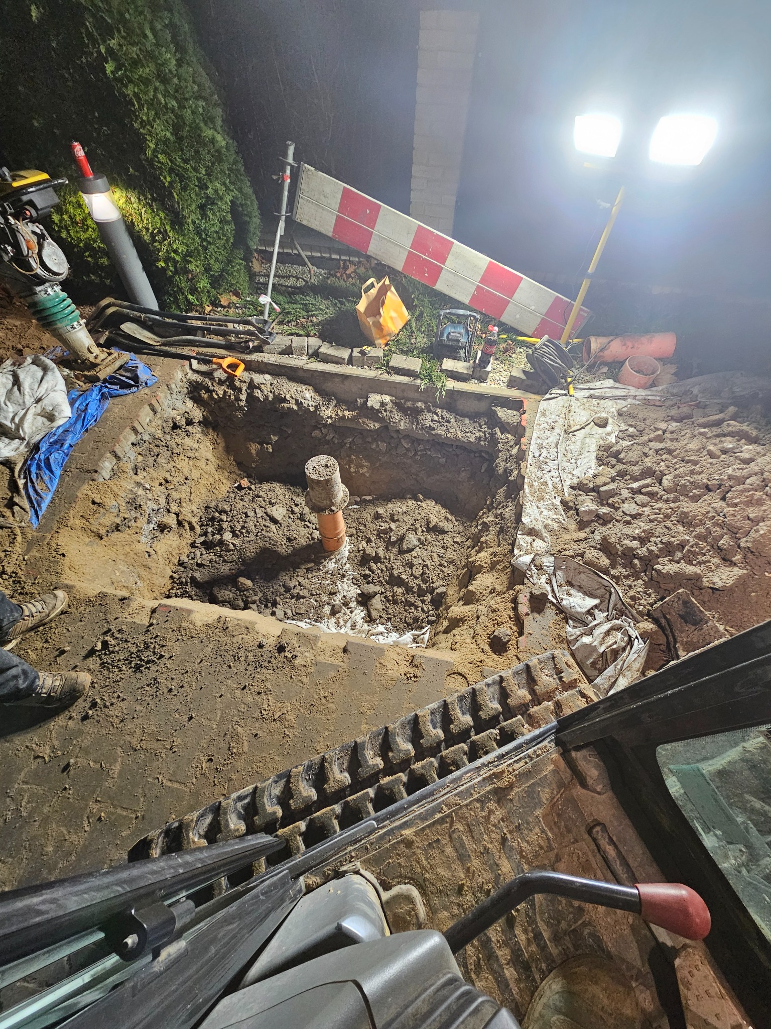 Głęboki wykop oświetlony lampą budowlaną, widok z kabiny koparki na pomarańczową rurę wystającą z ziemi, ubijarka wibracyjna i narzędzia obok wykopu.