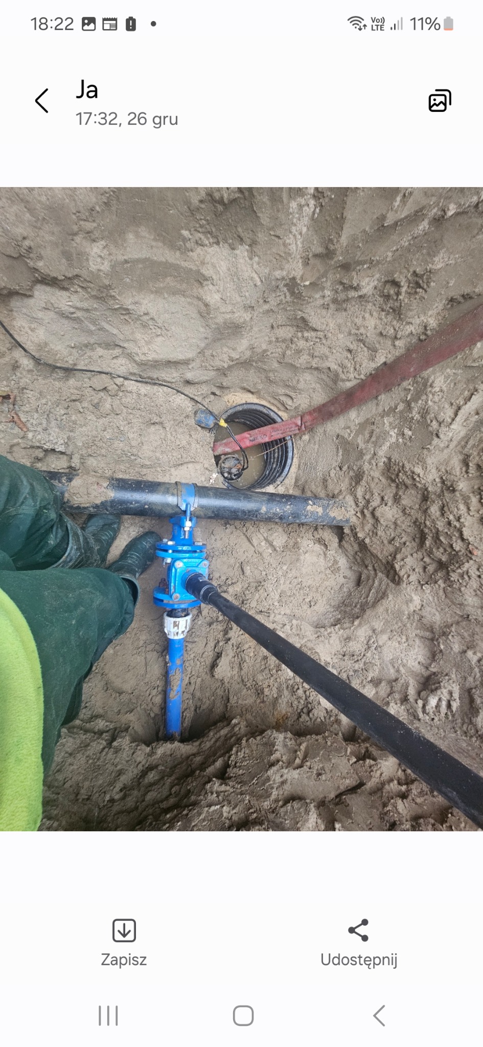 Wykop z widocznymi rurami wodociągowymi, zaworem i elementami podłączeniowymi, częściowo zasypany piaskiem, z elementami roboczymi w tle.