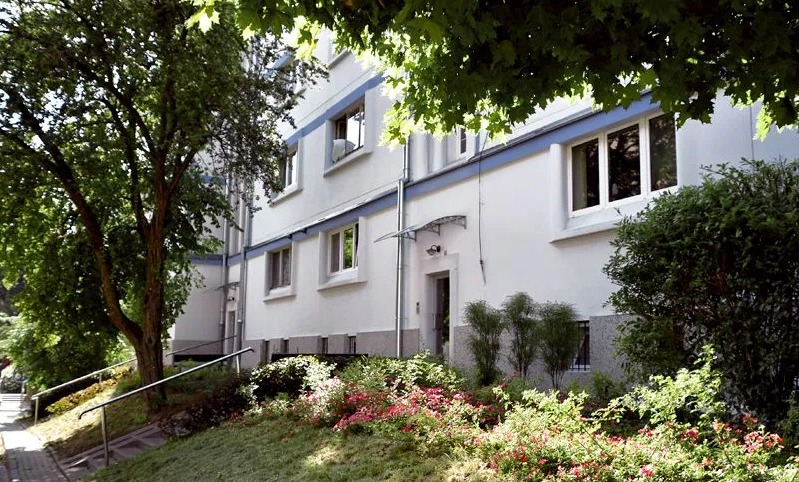 Elewacja trzypiętrowego budynku mieszkalnego z białymi ścianami i niebieskimi akcentami, widok z poziomu ogrodu z kwitnącymi krzewami.