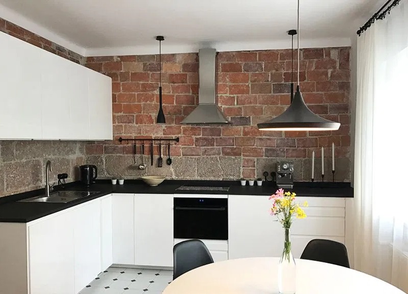 Nowoczesna kuchnia z białymi szafkami, czarnym blatem i ceglaną ścianą w tle, oświetlona wiszącymi lampami w stylu industrialnym, z kwiatami w wazonie na okrągłym stole.