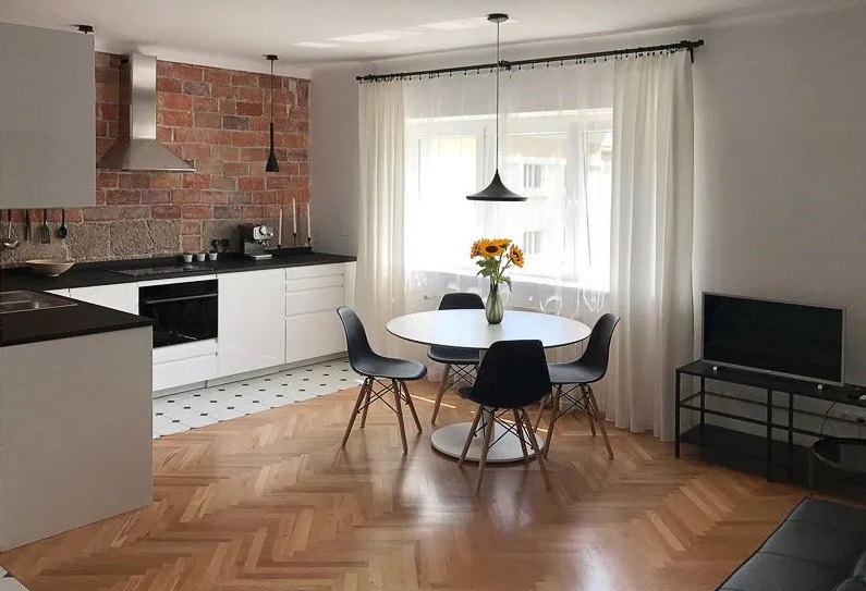 Jasne wnętrze mieszkania z ceglaną ścianą, białymi meblami kuchennymi, okrągłym stołem z bukietem słoneczników i parkietem w jodełkę.