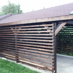 Drewniana konstrukcja z poziomych listew i spadzistym dachem z brązowej dachówki bitumicznej, widoczna z zewnątrz, na tle zieleni ogrodu i kostki brukowej.