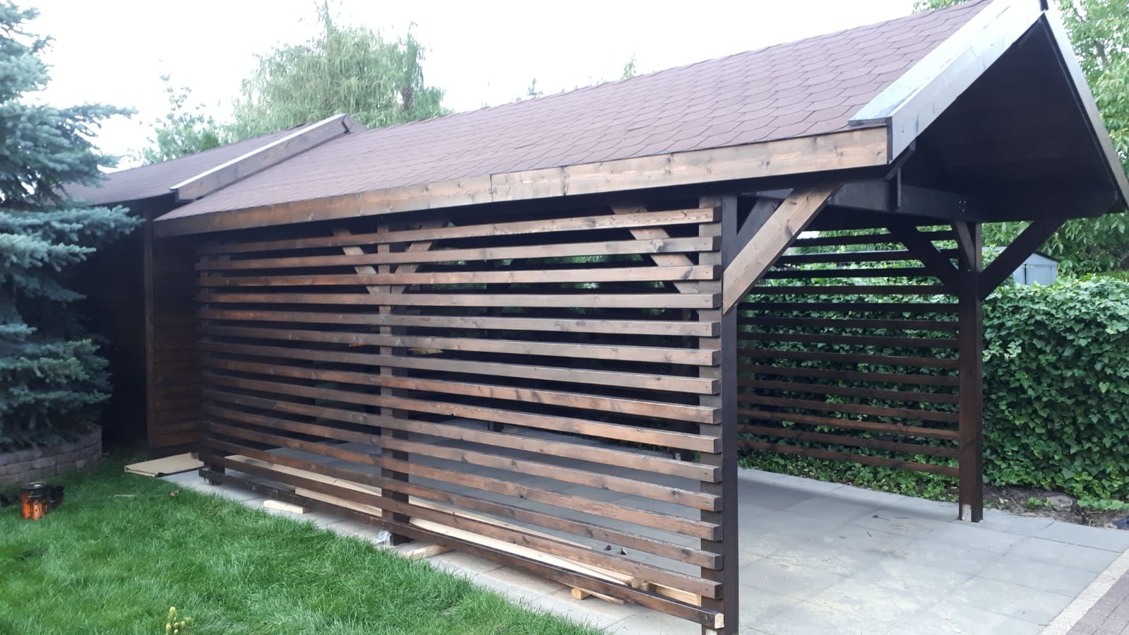 Drewniana konstrukcja z poziomych listew i spadzistym dachem z brązowej dachówki bitumicznej, widoczna z zewnątrz, na tle zieleni ogrodu i kostki brukowej.