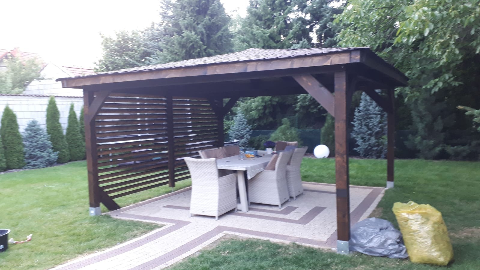 Zadaszona altana ogrodowa z drewnianą konstrukcją i lamelową ścianą, wyposażona w stół i krzesła, ustawiona na wybrukowanym patio, otoczona zielonym trawnikiem i drzewami.