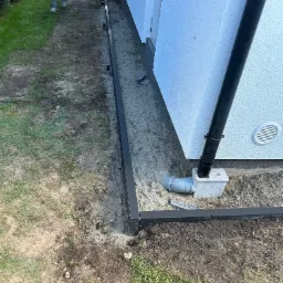 Przygotowanie podłoża pod ścieżkę z obrzeżami z tworzywa sztucznego, widoczny fragment trawnika, rynna spustowa i fragment elewacji budynku.