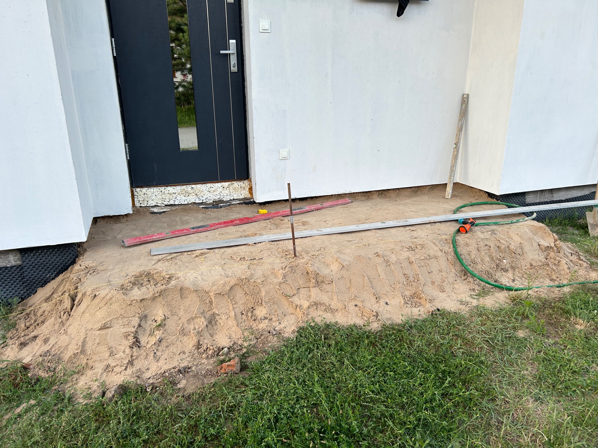Przygotowanie terenu przed wejściem do domu, widoczna warstwa piasku, poziomice, pręt, wąż ogrodowy, budynek z białą elewacją i ciemnymi drzwiami wejściowymi.
