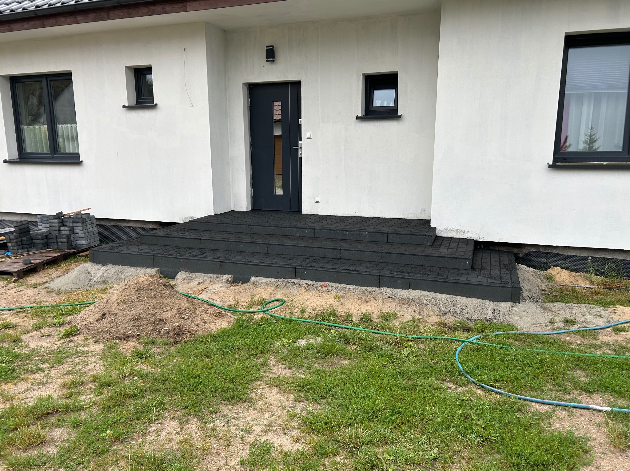 Nowo ułożone, ciemnoszare schody z kostki brukowej prowadzące do drzwi wejściowych nowoczesnego domu z białymi ścianami, widoczny stos kostki brukowej i węże ogrodowe na trawniku.