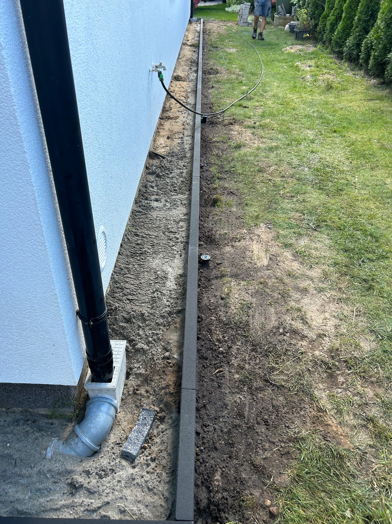 Przygotowanie terenu pod kostkę brukową: wyrównana ziemia wzdłuż ściany budynku z widocznym odpływem rynnowym i obrzeżem z betonowych krawężników, w tle trawnik i osoba.