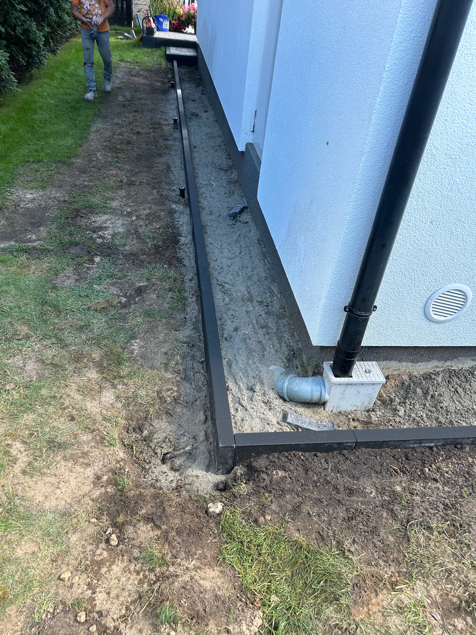 Przygotowanie podłoża pod ścieżkę z obrzeżami z tworzywa sztucznego, widoczny fragment trawnika, rynna spustowa i fragment elewacji budynku.