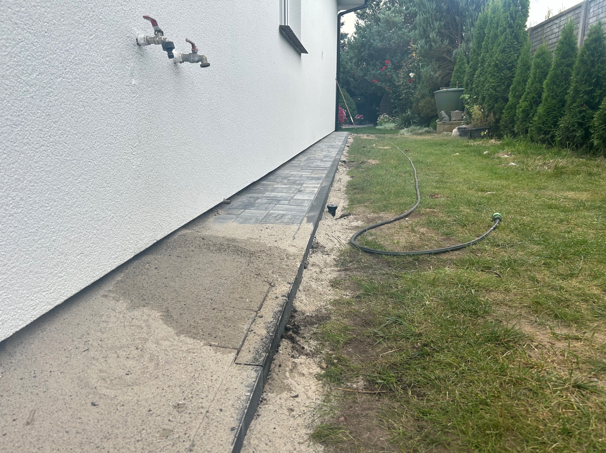 Ułożona kostka brukowa wzdłuż ściany domu, widoczne przyłącza wody na elewacji, trawnik z wężem ogrodowym w tle.