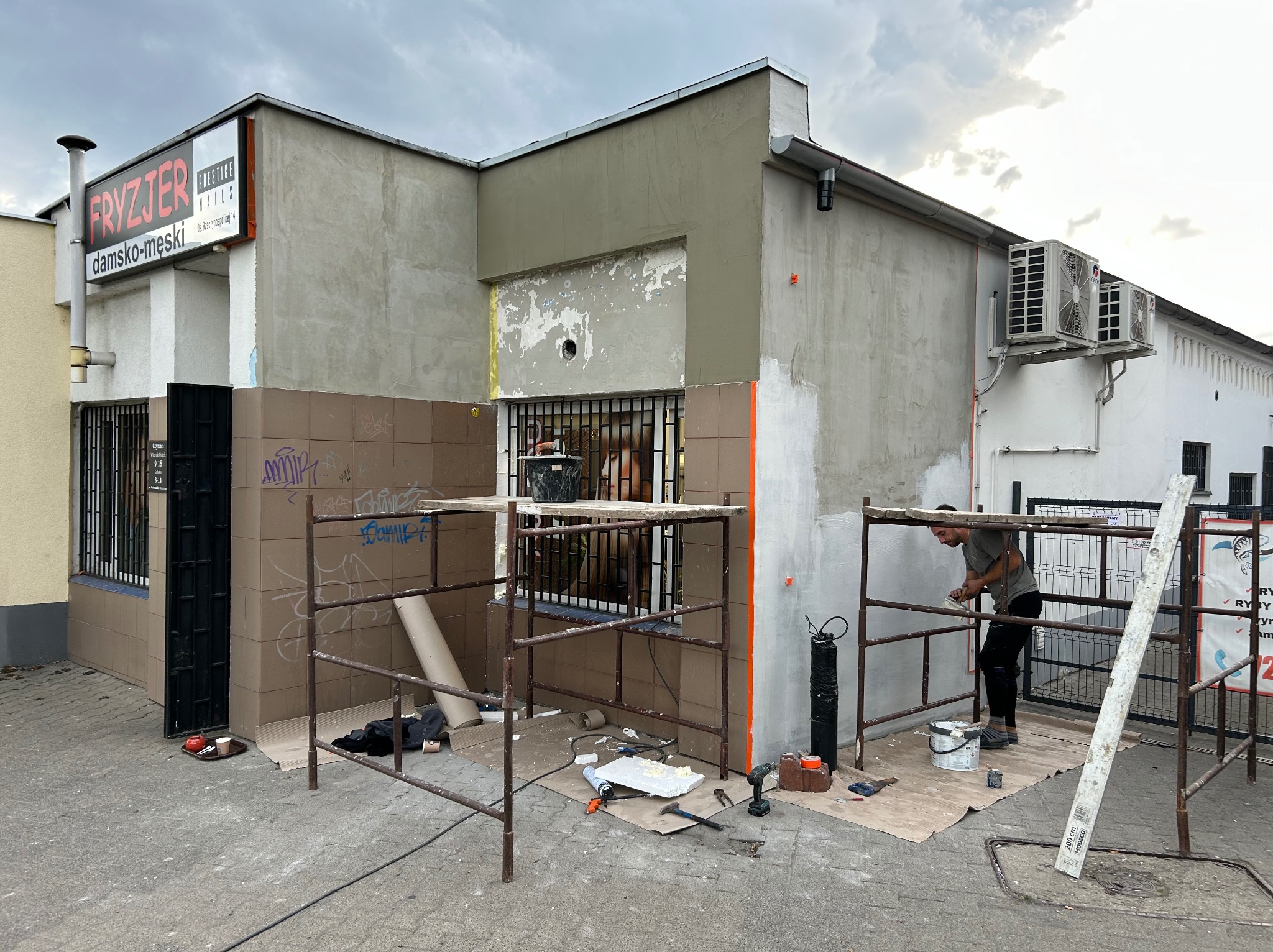 Pracownik na rusztowaniu maluje elewację budynku z szyldem 'Fryzjer damsko-męski', widoczne narzędzia i materiały budowlane, częściowo odnowiona fasada.