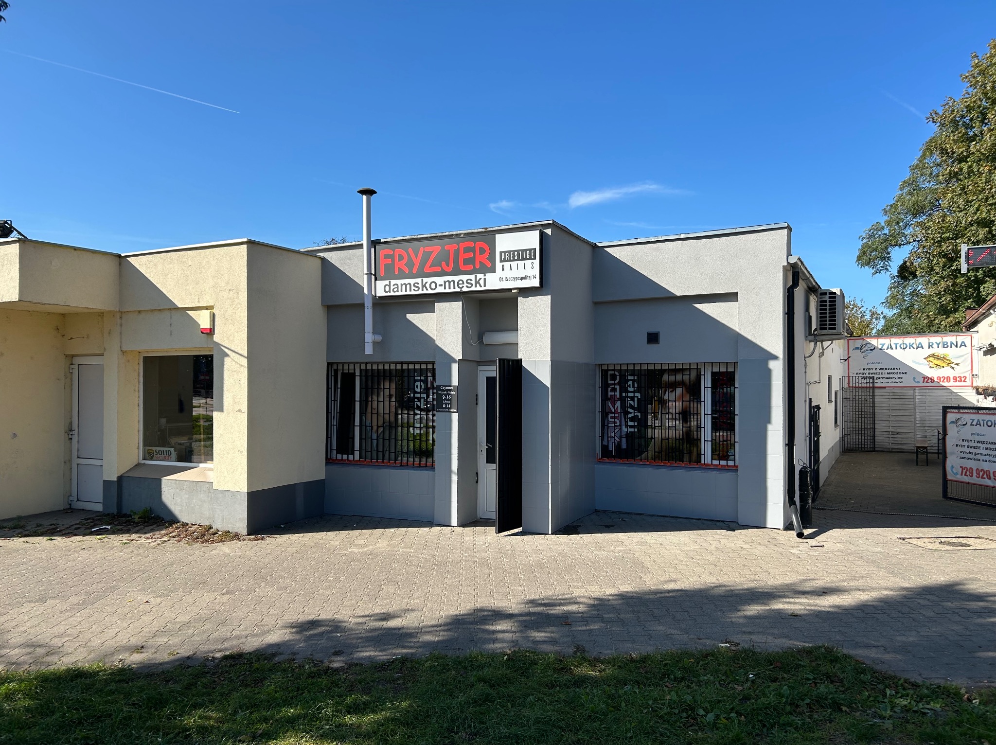 Fasada budynku z szyldem fryzjera damsko-męskiego 'Fryzjer Prestige Nails' z widocznym wejściem i oknami z kratami, obok budynek z reklamą 'Zatoka Rybna'.