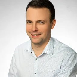 Portret uśmiechniętego mężczyzny w jasnej koszuli z drobnym wzorem na jednolitym, jasnym tle.