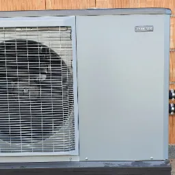 Szara jednostka zewnętrzna pompy ciepła NIBE F2040 zamontowana przy ścianie z czerwonej cegły dziurawki, z widocznymi dwoma czarnymi rurami z obejmami.