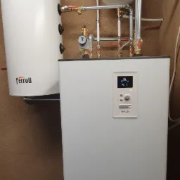 Biały, nowoczesny piec grzewczy Ferroli SHK 200 z podłączonymi rurami, manometrem i zbiornikiem wyrównawczym na tle ściany o fakturze korka. Na podłodze widoczne ślady po remoncie.