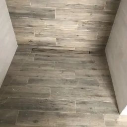 Wykończona podłoga w kabinie prysznicowej z odpływem liniowym, płytki imitujące drewno ułożone poziomo.