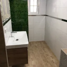 Nowoczesna łazienka z umywalką nablatową na szafce z drewnopodobnym frontem, białe płytki z delikatnym wzorem na ścianach, podłoga wyłożona płytkami imitującymi drewno, ściana prysznicowa...