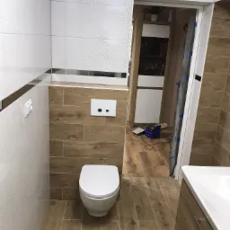 Nowoczesna toaleta podwieszana w łazience wykończonej płytkami imitującymi drewno, z widoczną częścią umywalki i otwartymi drzwiami do dalszej części mieszkania, gdzie widać karton i narzędzia.