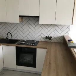 Nowoczesna kuchnia z białymi szafkami, drewnianym blatem i czarnym zlewem oraz płytą grzewczą, wykończona białymi płytkami ułożonymi w jodełkę, widoczne gniazdka elektryczne i akcesoria pomiarowe.