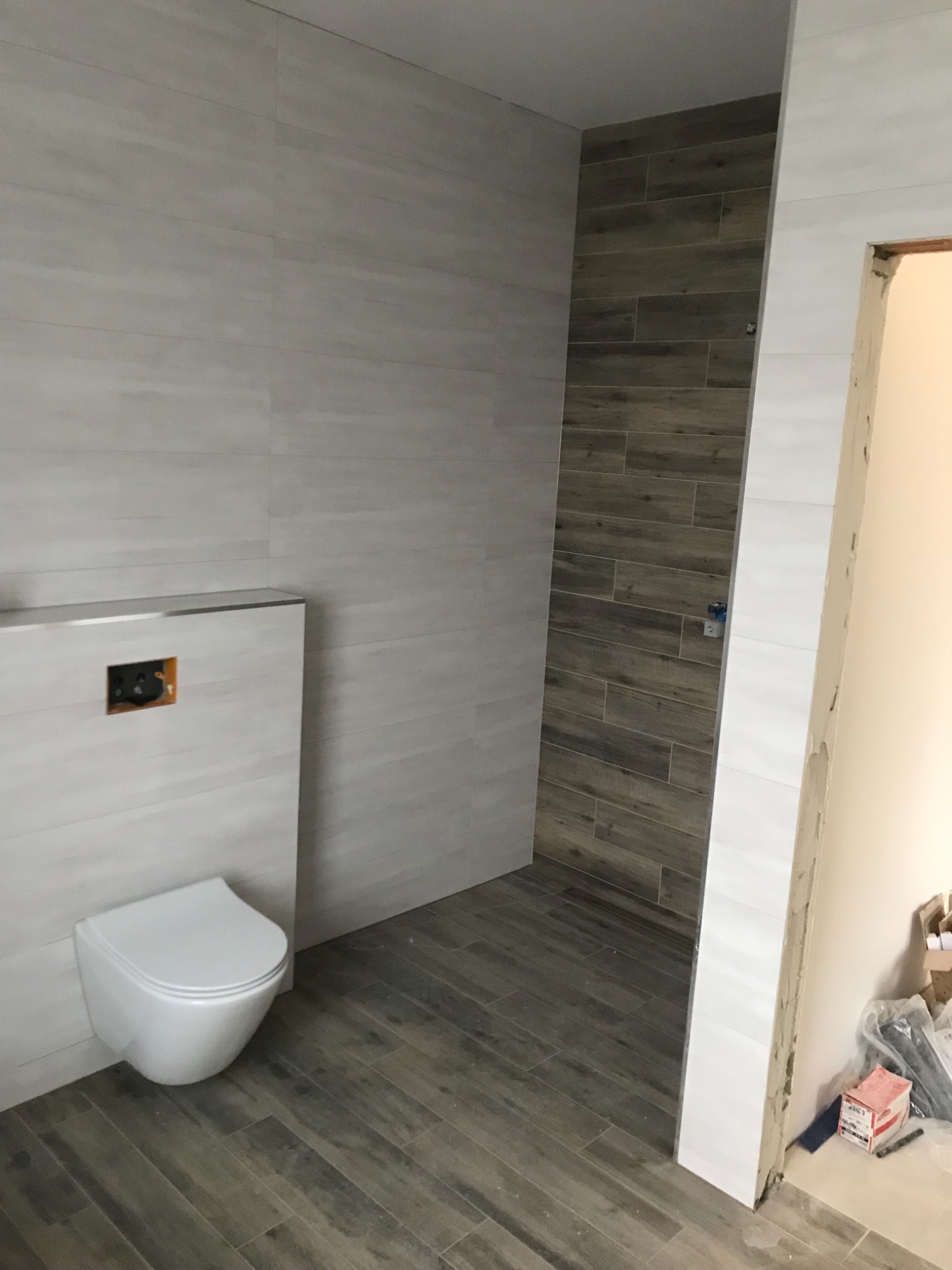 Świeżo wykafelkowana łazienka z podwieszaną toaletą, jasnoszare płytki na ścianach i podłodze imitujące drewno, z akcentem ciemniejszych płytek w strefie prysznica, widoczny otwór na przyciski...