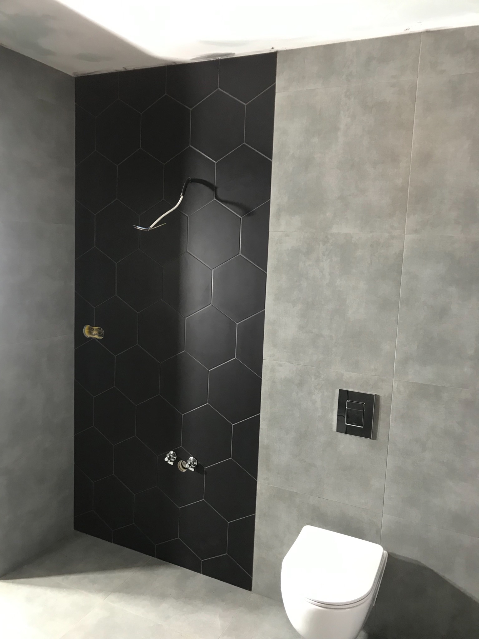 Łazienka w trakcie wykańczania: czarne, heksagonalne płytki na ścianie kontrastują z szarymi, wielkoformatowymi płytami; widoczne wyprowadzenia elektryczne i hydrauliczne, częściowo zamontowana...