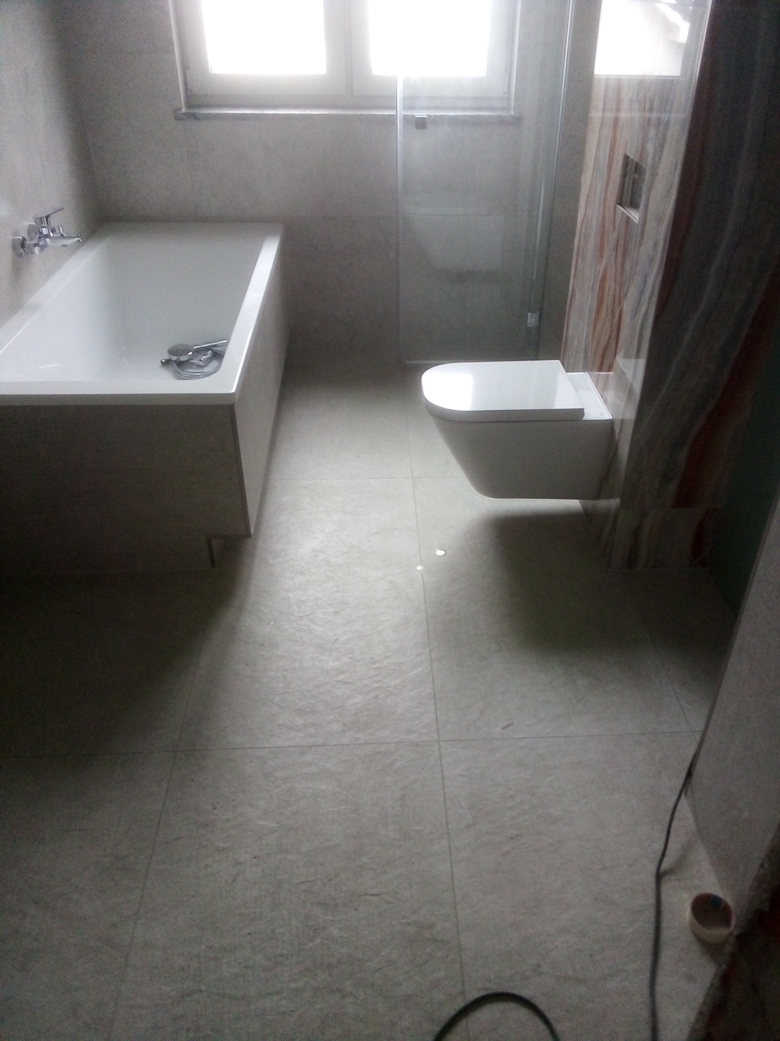 Nowoczesna łazienka z białą wanną, podwieszaną toaletą i beżowymi płytkami podłogowymi oraz częściowo marmurowymi płytkami ściennymi pod prysznicem.