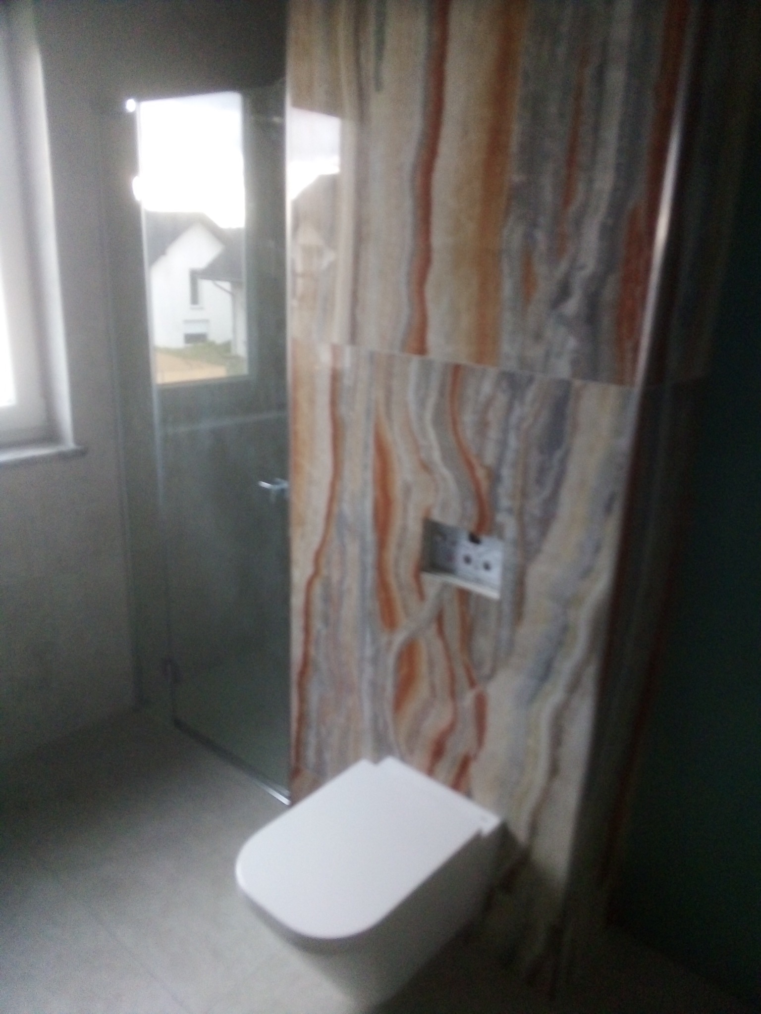 Nowoczesna łazienka w trakcie wykańczania: widoczna toaleta wisząca, ściana wyłożona płytkami imitującymi marmur z zamontowanym panelem sterującym spłuczką, oraz szklana kabina prysznicowa. W tle...