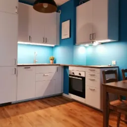 Nowoczesna kuchnia z białymi szafkami, błękitną ścianą, drewnianą podłogą i czarną lampą wiszącą. Na blacie stoi doniczka z rośliną, a na ścianie wisi obrazek z motywem kota.