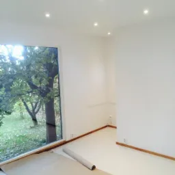 Białe, świeżo pomalowane pomieszczenie z dużym oknem, zabezpieczone kartonem falistym na podłodze, widoczne punktowe oświetlenie sufitowe.