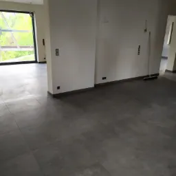 Wnętrze w stanie deweloperskim z szarą, matową podłogą z płytek, widoczne gniazdka elektryczne na białych ścianach i szczotka do zamiatania oparta o ścianę.