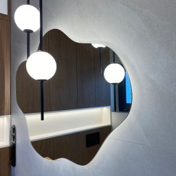 Nie Do Zbicia - Nieregularny kształt szklanego lustra z podświetleniem LED, odbijającego fragment drewnianej zabudowy meblowej i wiszące lampy kuliste, zamontowanego na ścianie z szarego kamienia.