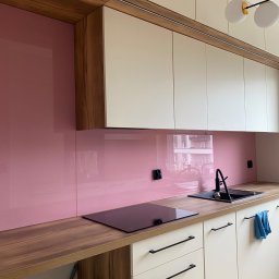 Nie Do Zbicia - Nowoczesna kuchnia z różowym panelem szklanym nad blatem, drewnianym blatem i białymi szafkami. Widoczny zlew, płyta indukcyjna i minimalistyczne uchwyty.