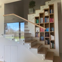 Nie Do Zbicia - Nowoczesne schody z drewnianymi stopniami i szklaną balustradą, wspornikami punktowymi. Obok schodów regał na książki wbudowany w ścianę.