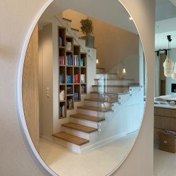 Nie Do Zbicia - Okrągłe lustro odbija nowoczesne schody z drewnianymi stopniami i szklaną balustradą, obok wbudowany regał na książki. Jasne, minimalistyczne wnętrze.