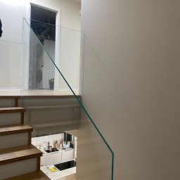 Nie Do Zbicia - Szklana balustrada na schodach z drewnianymi stopniami, widok z góry na kuchnię i pralkę w tle. Minimalistyczny design, jasne wnętrze.