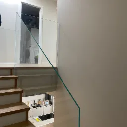 Szklana balustrada na schodach z drewnianymi stopniami, widok z góry na kuchnię i pralkę w tle. Minimalistyczny design, jasne wnętrze.