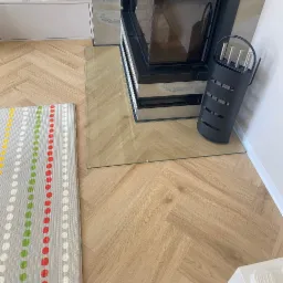 Szklana podłoga pod kominkiem, zabezpieczająca drewniany parkiet ułożony w jodełkę. Obok czarny stojak na narzędzia kominkowe i mata dla dzieci w kropki.