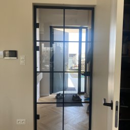 Nie Do Zbicia - Szklane drzwi w czarnej ramie, widok z wnętrza domu na zewnątrz. Podłoga z parkietem w jodełkę, w tle widoczne buty i wycieraczka.