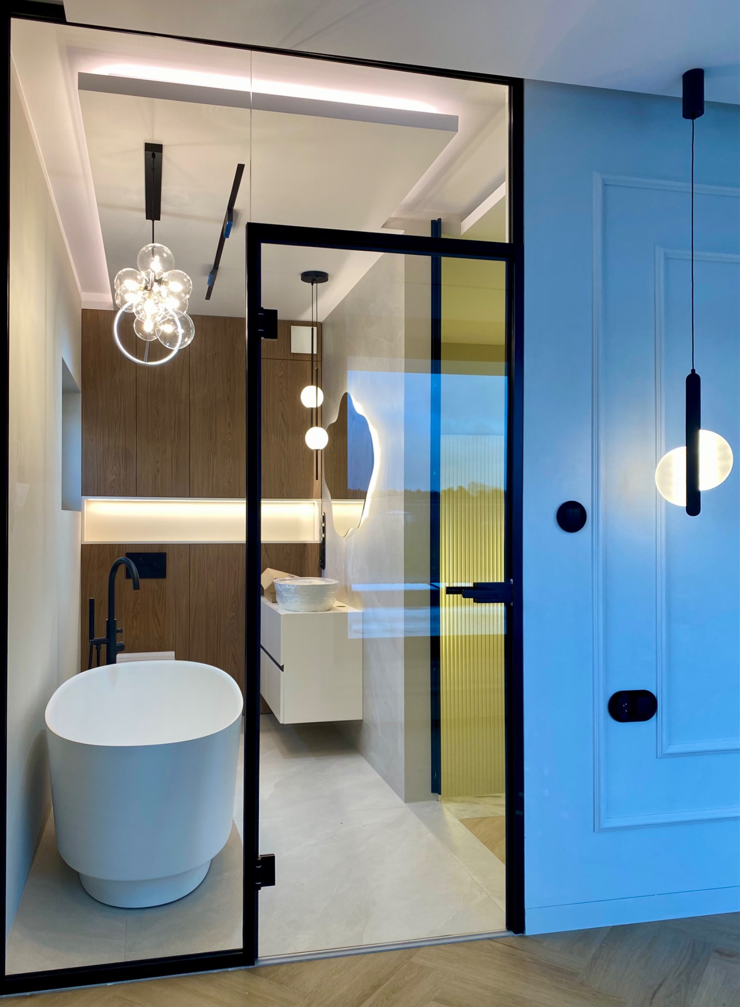 Nowoczesna łazienka z kabiną prysznicową oddzieloną szklanymi drzwiami z czarną ramą, widoczna wolnostojąca wanna i designerskie oświetlenie.